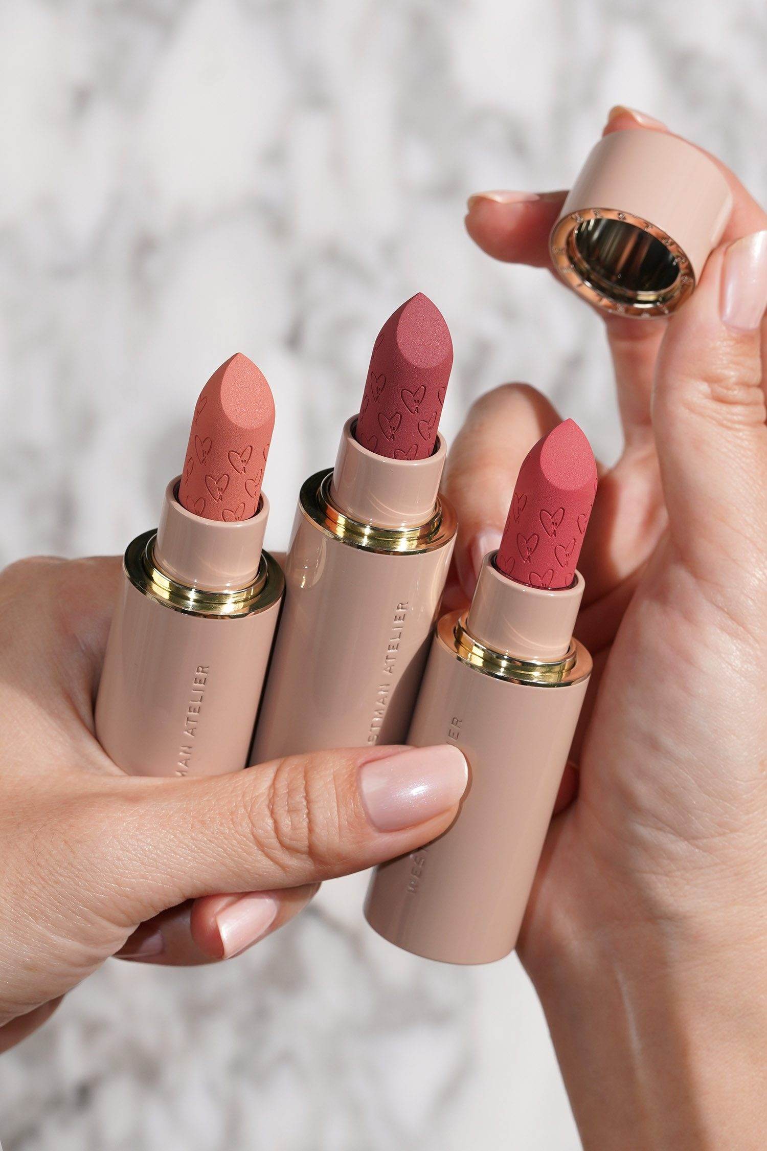 Close-up of three luxurious lipsticks in soft nude, mauve, and pink shades held by hands, showcasing LIPS collection