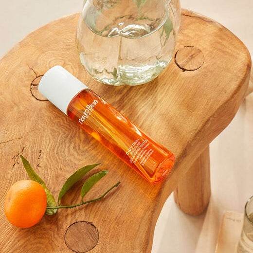 Natura Bisse Revitalizing Cleanser and Makeup Remover in an orange bottle on a wooden table with a tangerine