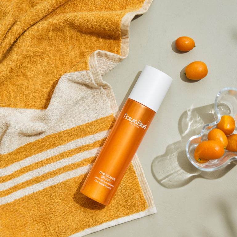 Natura Bisse Revitalizing Citrus Spray in an orange bottle next to a towel and fresh citrus fruits