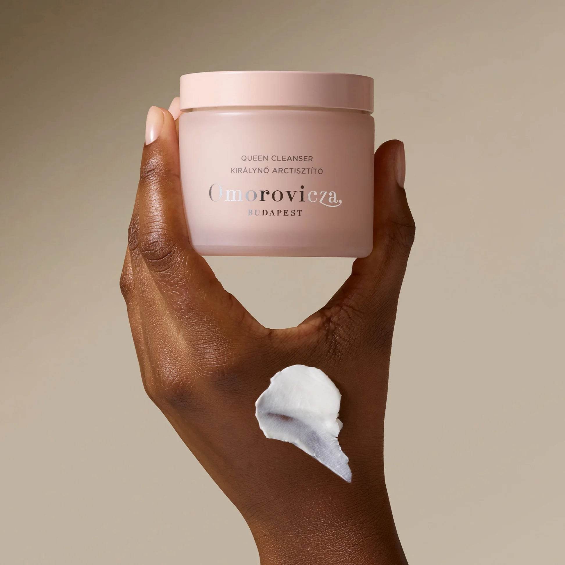 Omorovicza Queen Cream Cleanser in a pink jar held in hand, showcasing creamy texture on skin