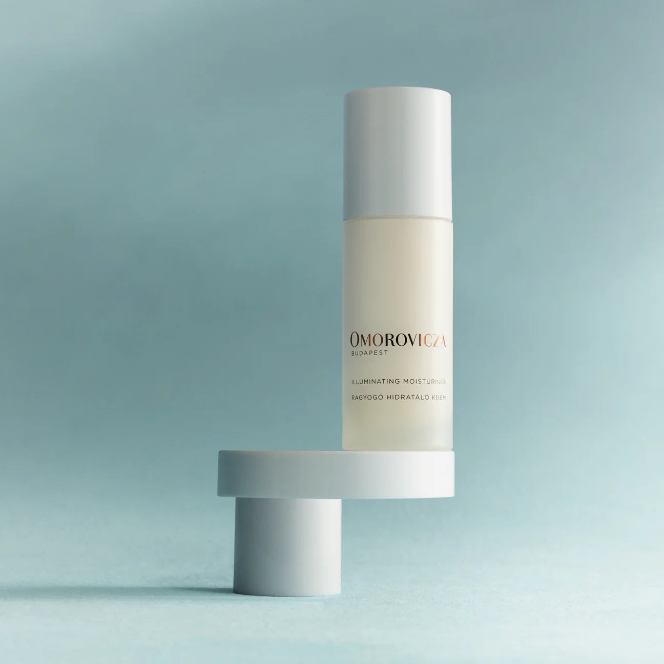 Omorovicza Illuminating Moisturizer bottle with white cap on minimalist pedestal against blue background