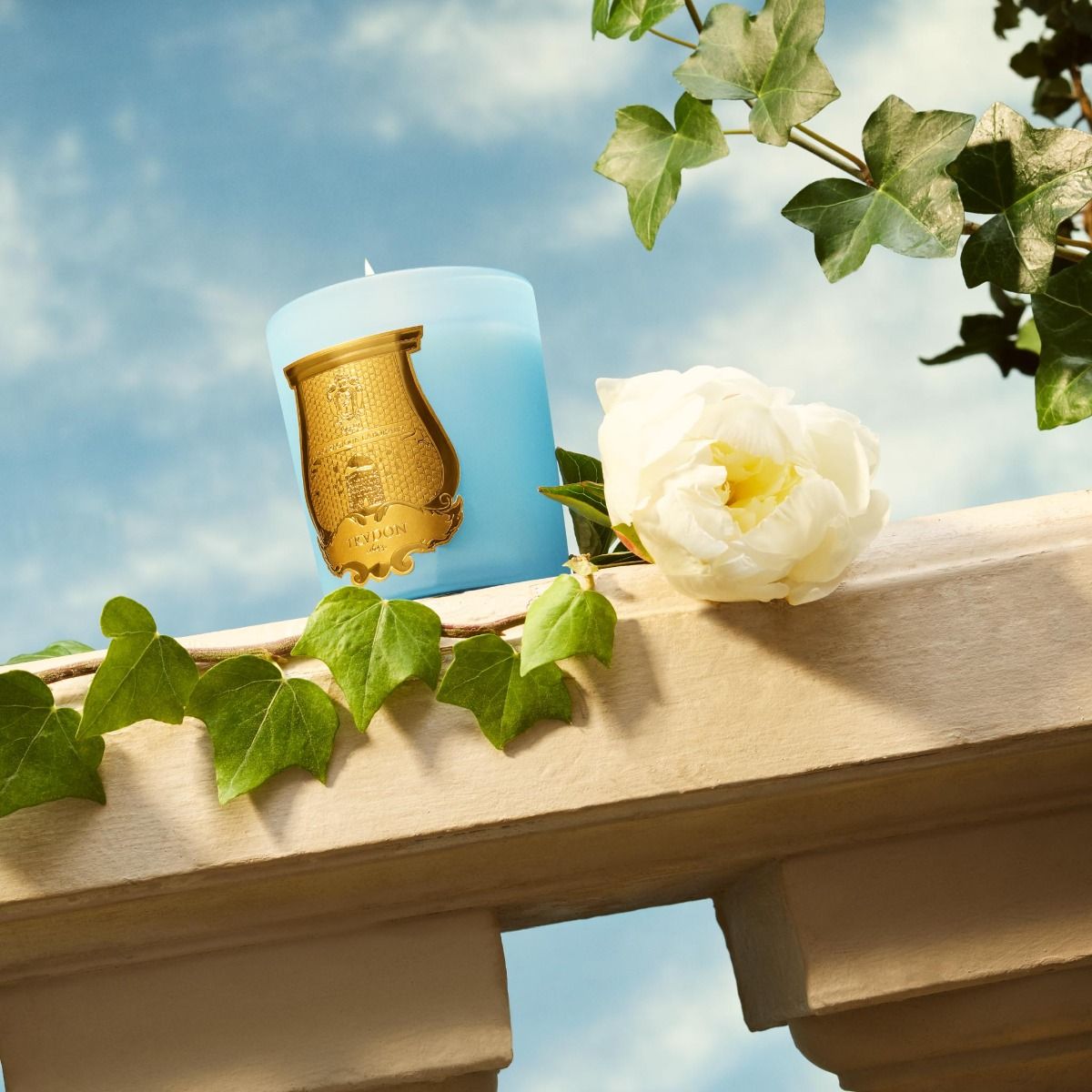 London Beauty Versailles Candle in blue with gold label beside white rose and green leaves on stone ledge