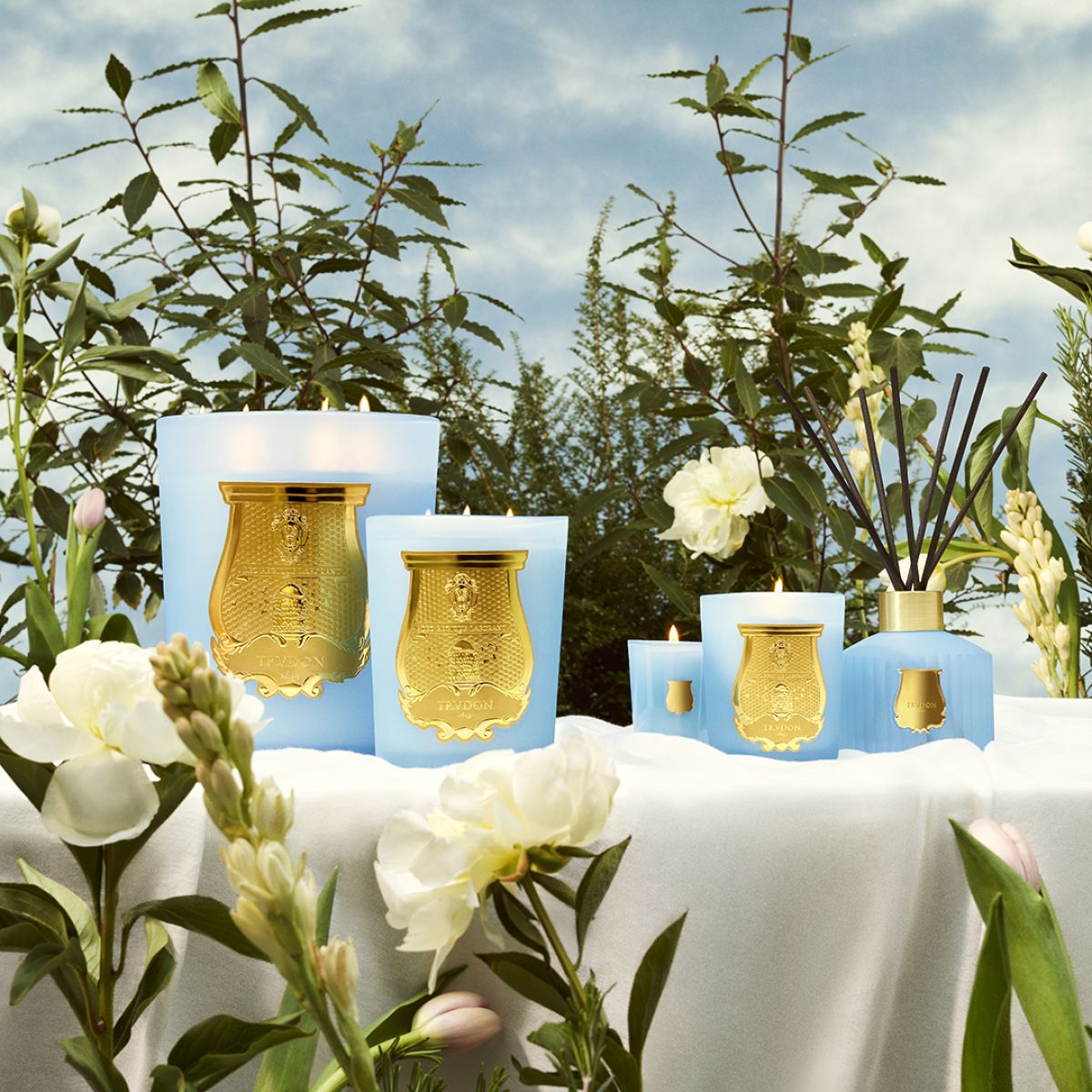 London Beauty Versailles Candle collection in elegant blue jars with gold labels surrounded by white flowers outdoors