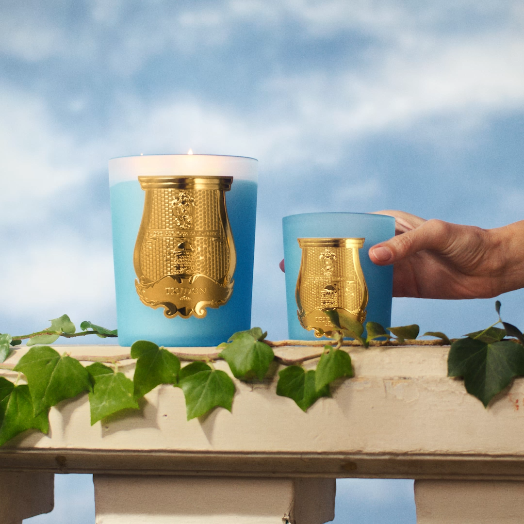 Two blue Versailles Candle jars with gold labels on a stone ledge, one lit and a hand holding the smaller jar